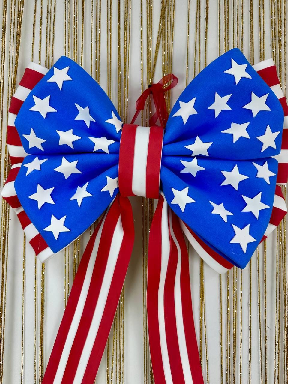May include: A large decorative bow featuring a patriotic design. The bow has a blue center adorned with white stars, red and white striped edges, and long red and white striped ribbons. A red ribbon is attached for hanging.