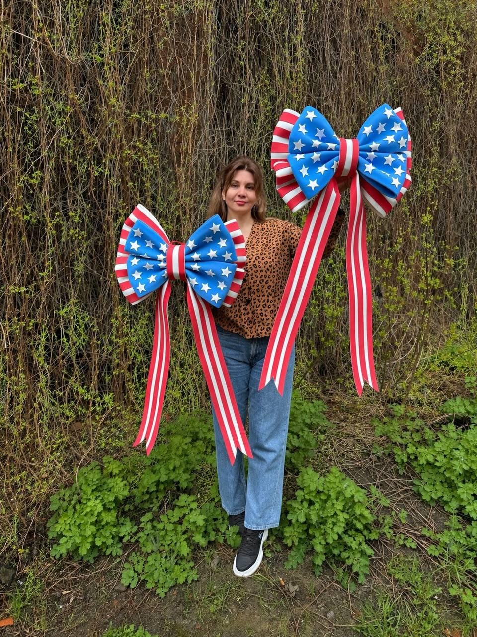 May include: Two large decorative bows with a patriotic theme. The bows feature a blue center with white stars and red and white striped edges and streamers. The bows are held up in front of a background of green foliage.