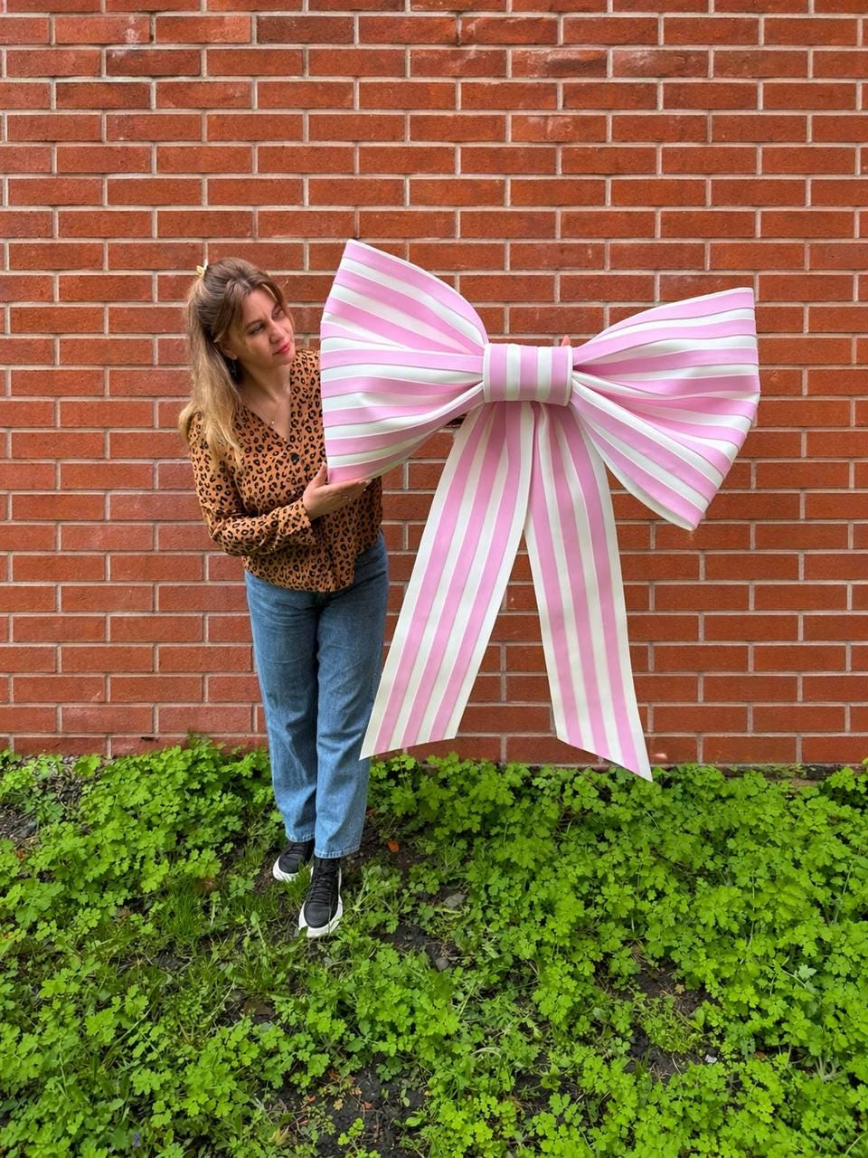 May include: A large, decorative bow with pink and white stripes. The bow is held in front of a red brick wall and green foliage. The bow's size suggests it could be used for a party or event decoration.