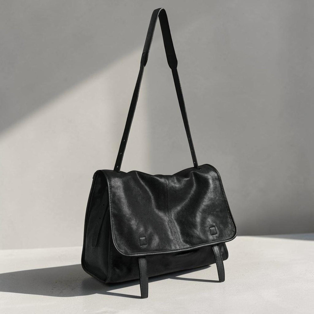 May include: A black leather messenger bag with a flap closure and adjustable shoulder strap. The bag has a rectangular shape with a soft, textured appearance. The strap is thin and the bag has a minimalist design.