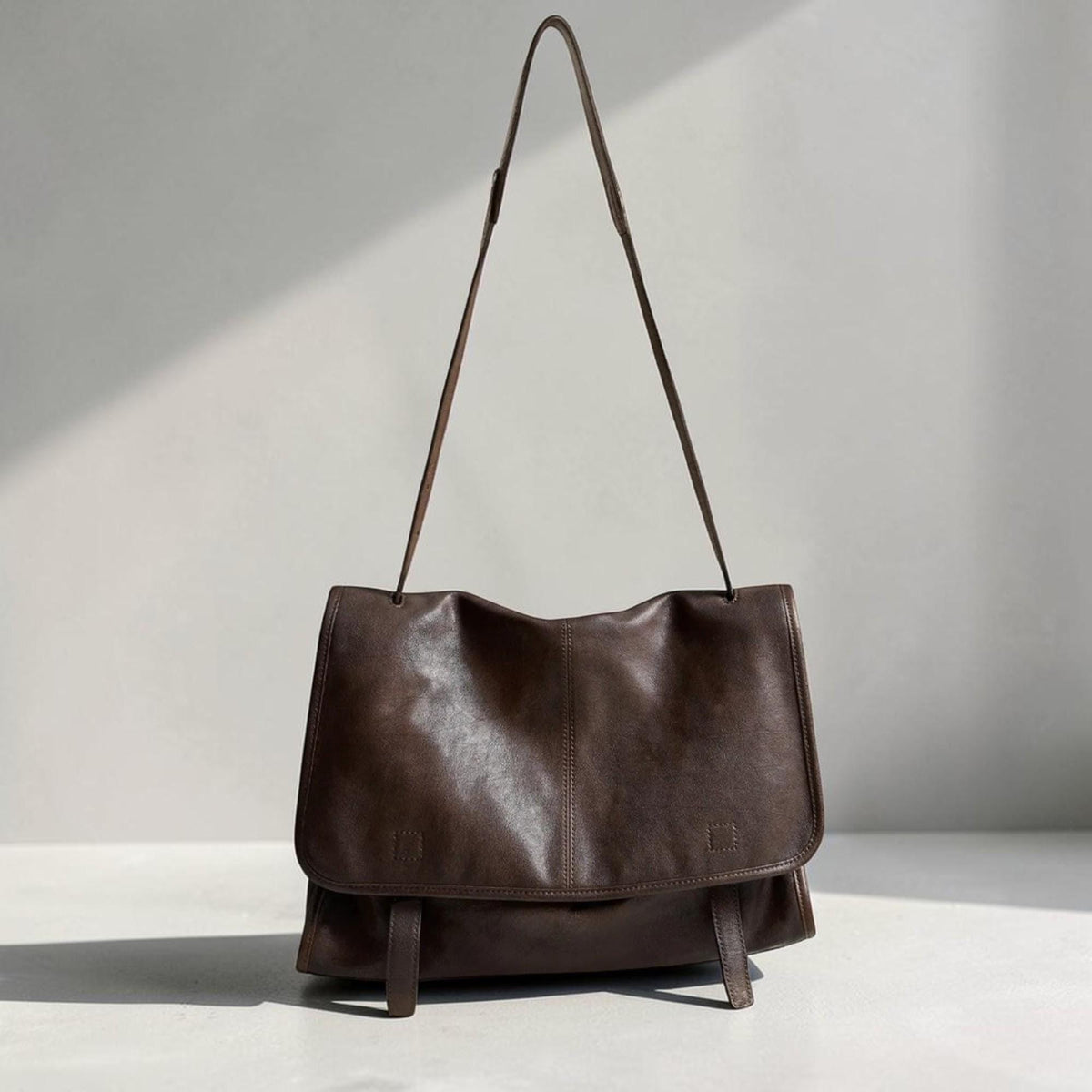 May include: A brown leather messenger bag with a long shoulder strap. The bag has a flap closure and two small buckles on the front. The leather appears to be soft and has a slightly wrinkled texture. The bag is designed to be carried over the shoulder.