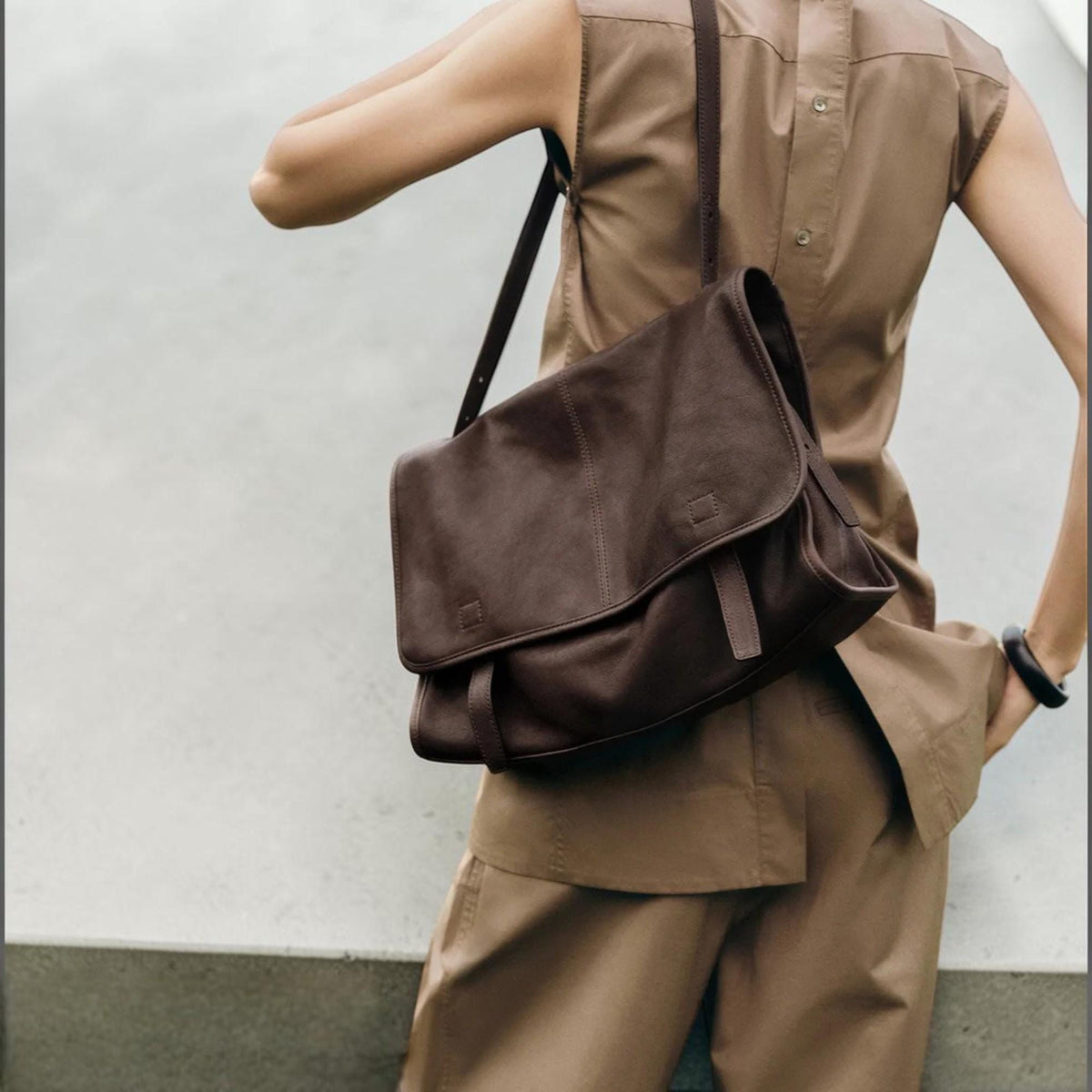 May include: A brown leather messenger bag with a flap closure and adjustable shoulder strap. The bag is worn over the shoulder and has a rectangular shape. The bag is made of a soft, textured leather.