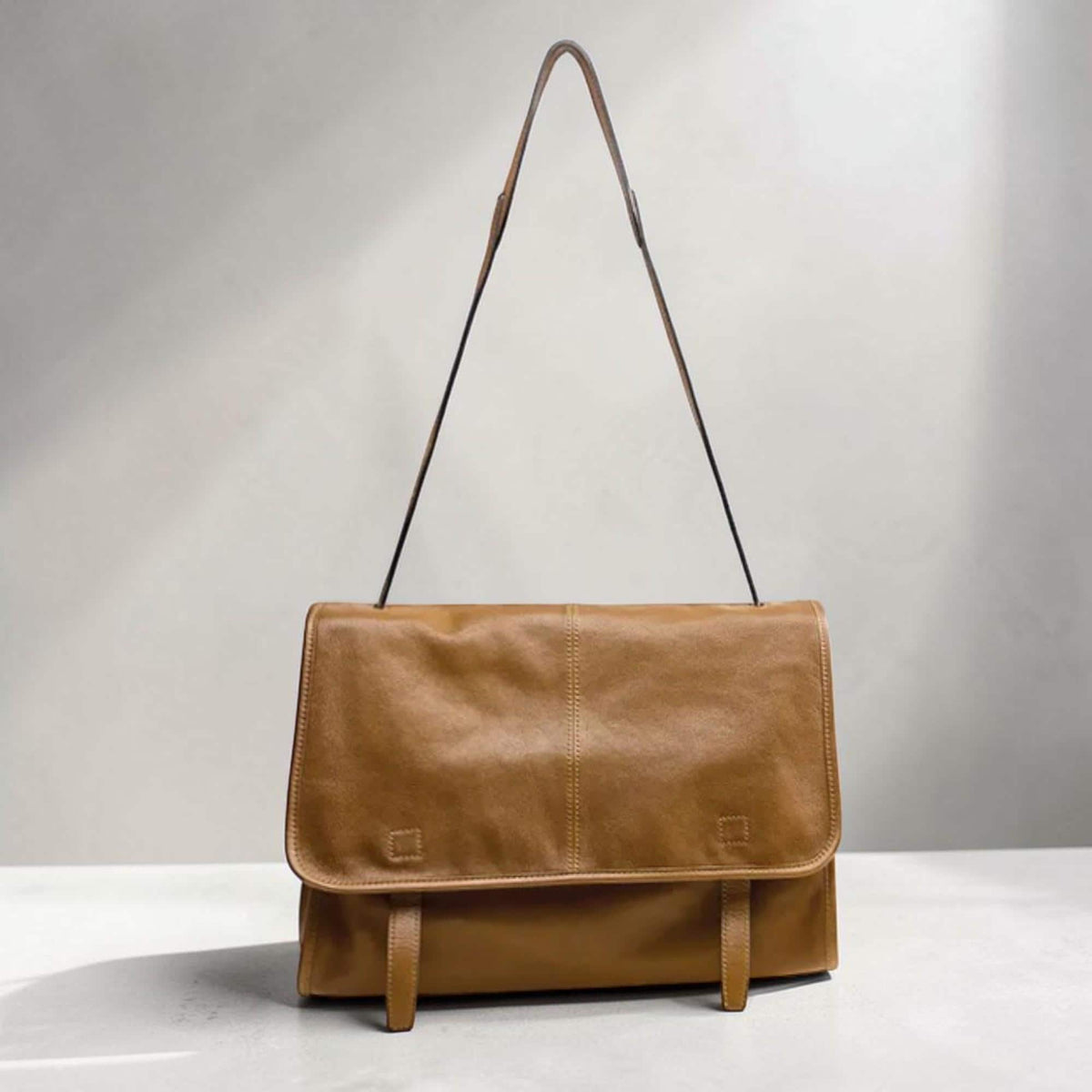 May include: A brown leather messenger bag with a long, thin strap. The bag has a rectangular shape with a flap closure and two buckle details. The leather has a smooth texture and a warm, natural color.