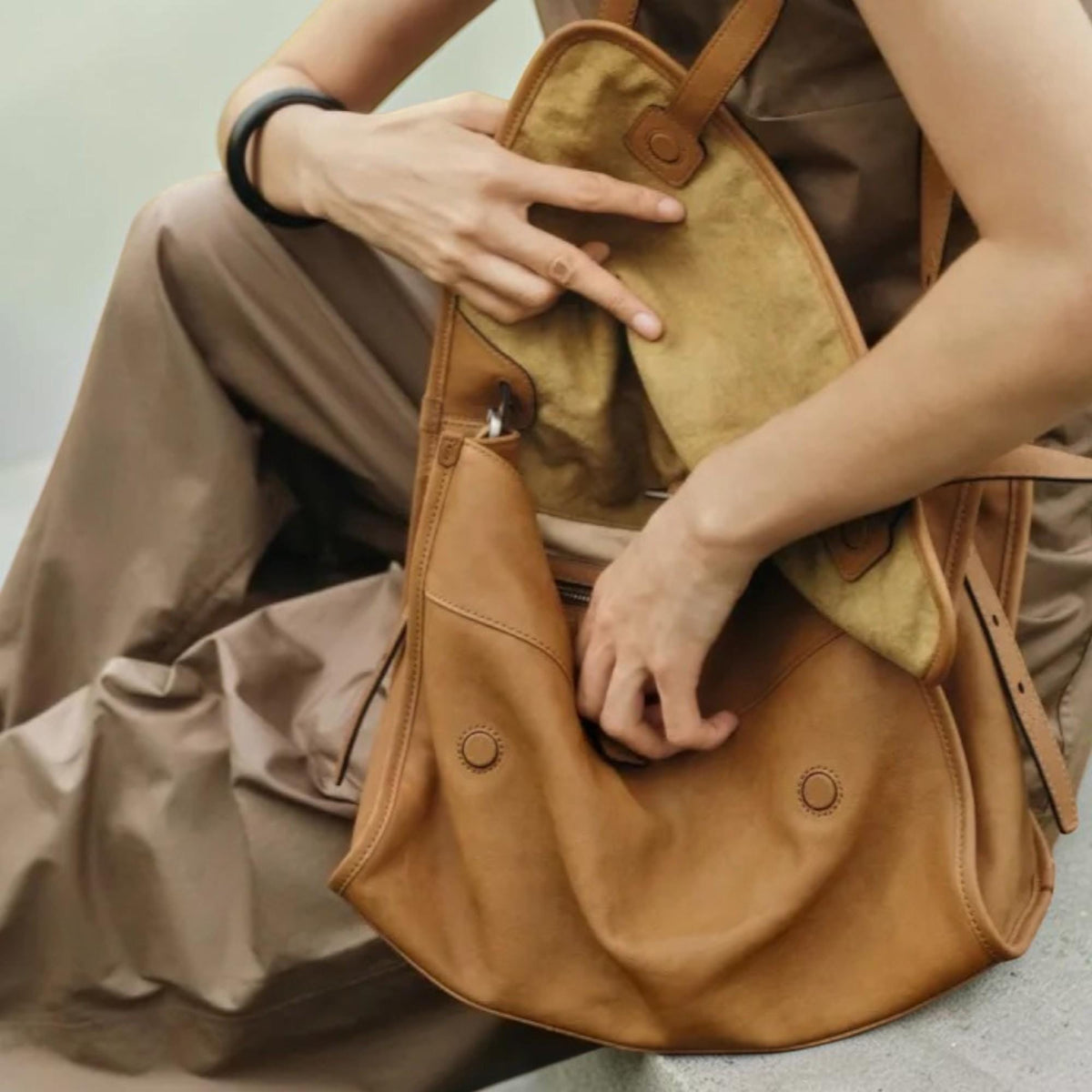 May include: A tan leather shoulder bag with a curved design. The bag has a flap closure and a long strap. The interior is a lighter tan color. Two circular rivets are visible on the front of the bag.