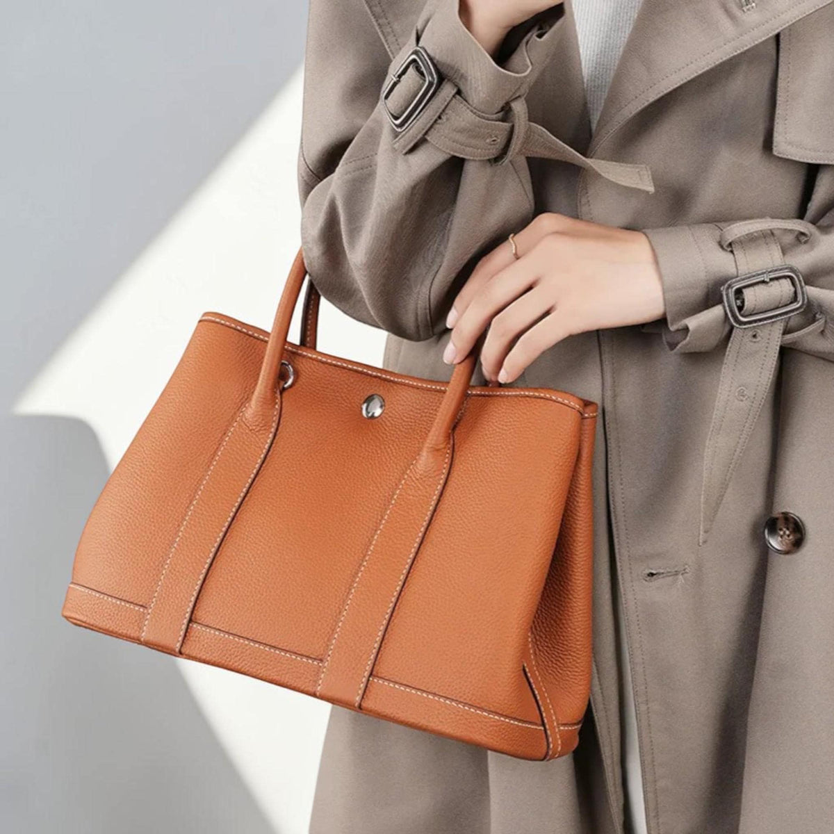 May include: A rectangular, tan leather handbag with two top handles and a silver clasp. The bag features white stitching along the seams and a structured design. The bag is held by a person wearing a tan trench coat.