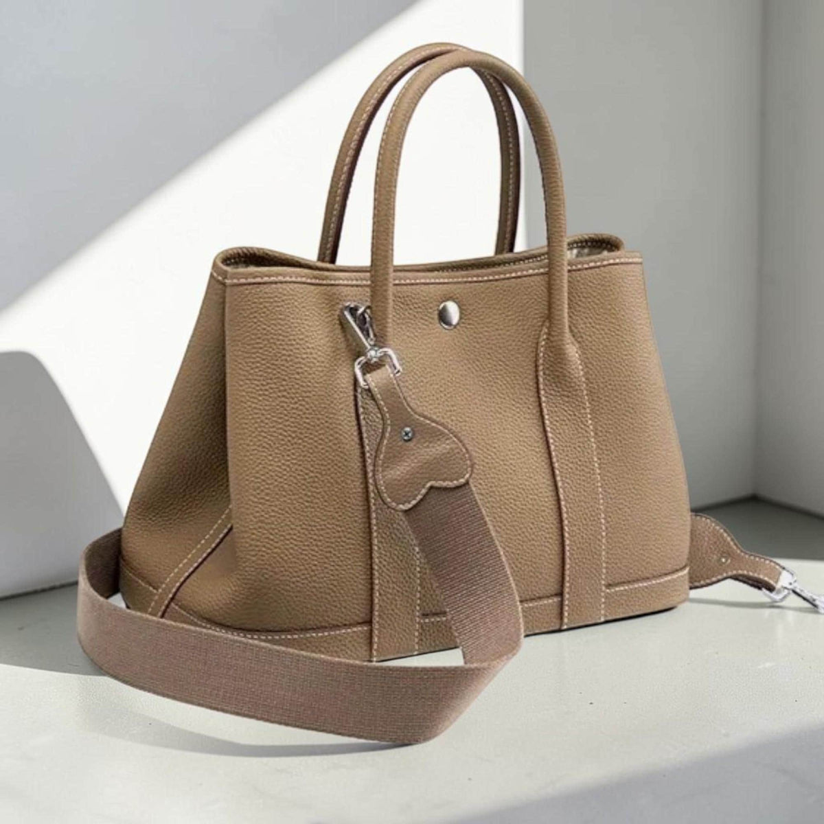 May include: A tan leather tote bag with a detachable shoulder strap. The bag has two handles and a silver-tone clasp. The stitching is a lighter color than the bag. The bag is sitting on a white surface.