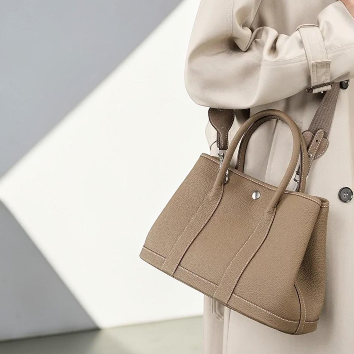 May include: A taupe-colored handbag with a top handle and a detachable shoulder strap. The bag features a structured design with visible stitching and a silver-tone closure. The bag is made of a textured material.
