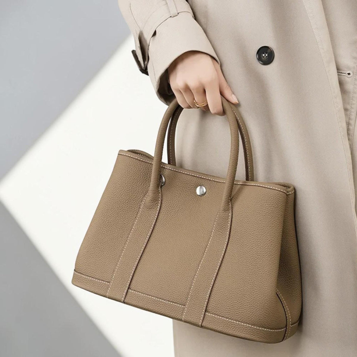 May include: A taupe-colored leather tote bag with two rounded handles and a structured design. The bag features white stitching along the seams and a silver-toned metal closure. The bag is held by a person wearing a beige trench coat.