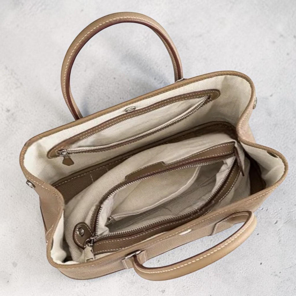 May include: A tan leather tote bag with two top handles and a structured design. The interior features multiple compartments with zippered closures and a light-colored lining. The bag has a classic, elegant style.