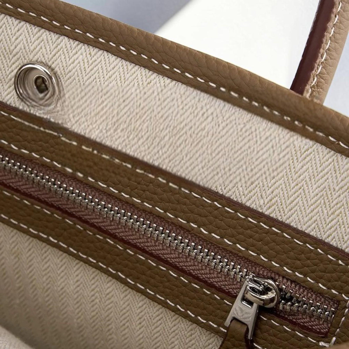 May include: Close-up of a brown leather tote bag interior. The bag features a beige herringbone fabric lining, a silver zipper, and white stitching details. The leather has a textured appearance.