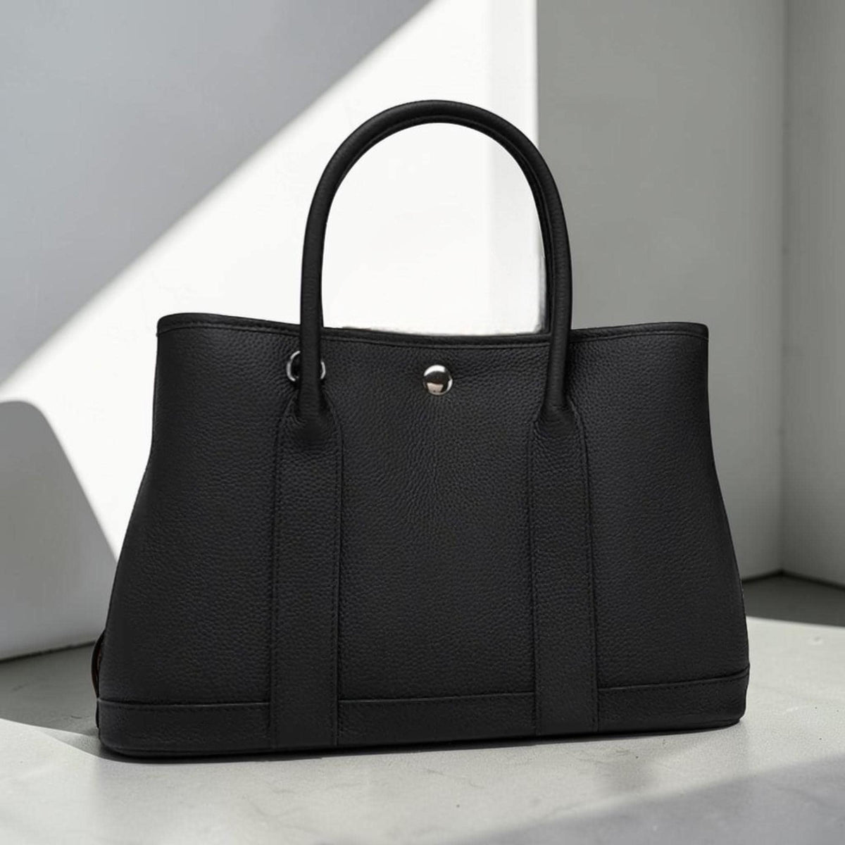 May include: A black tote bag with a structured design. The bag features a top handle and two smaller handles on the front. The bag has a silver metal clasp and is made of a textured material.