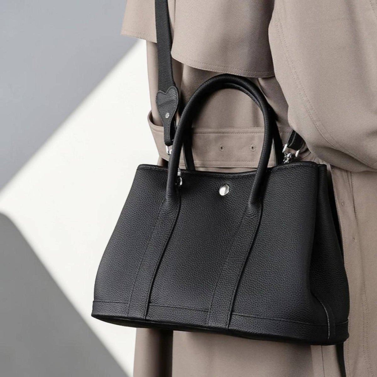 May include: A black leather handbag with a structured design. The bag features two top handles and a detachable shoulder strap. A silver-toned button detail is visible on the front. The bag is carried by a person wearing a tan trench coat.