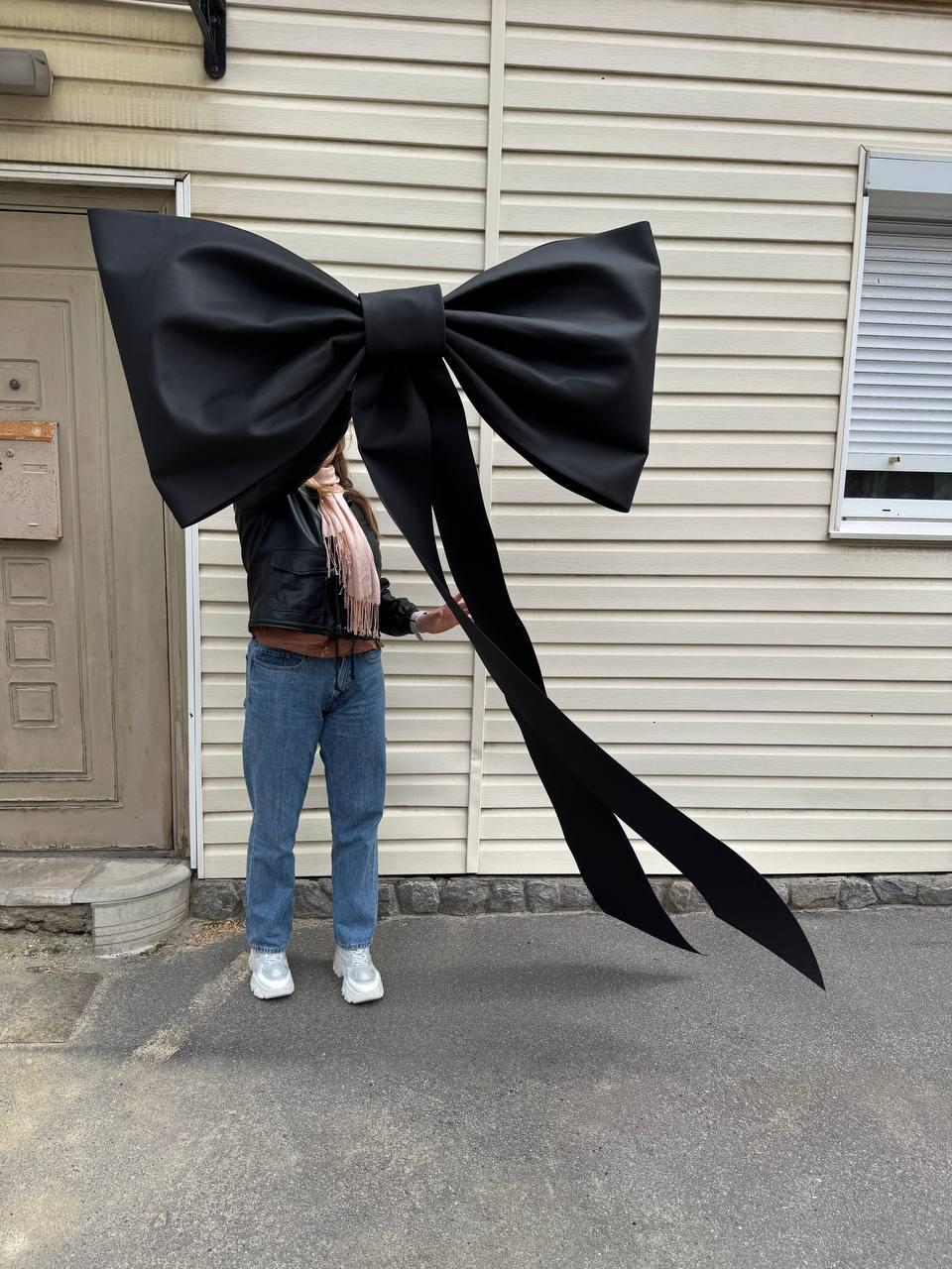 May include: A large, black bow with long, flowing ribbons. The bow is made of a shiny fabric and is held up in front of a person. The bow is a statement piece, perfect for a party or special event.
