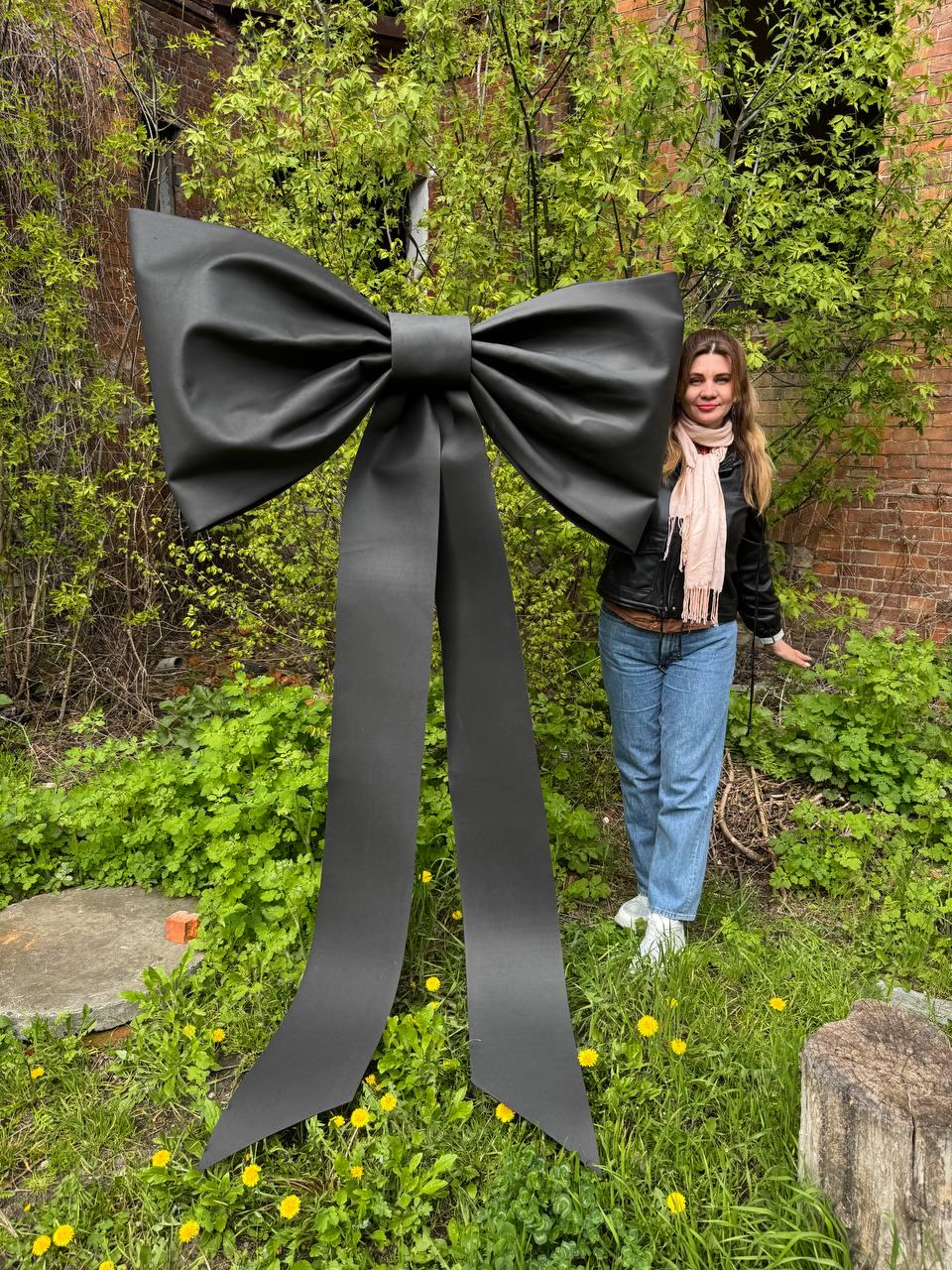 May include: A large, black bow stands in a grassy area with a person standing next to it. The bow is made of a smooth, satin-like material and has long, flowing tails. The background includes green foliage and a brick building.