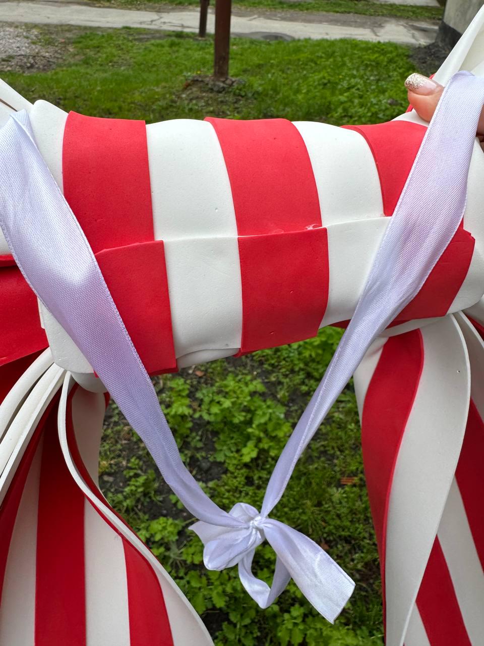 May include: A large, candy-shaped decoration with alternating red and white stripes. White ribbon is tied in a bow around the center. The decoration is outdoors, with green grass in the background.