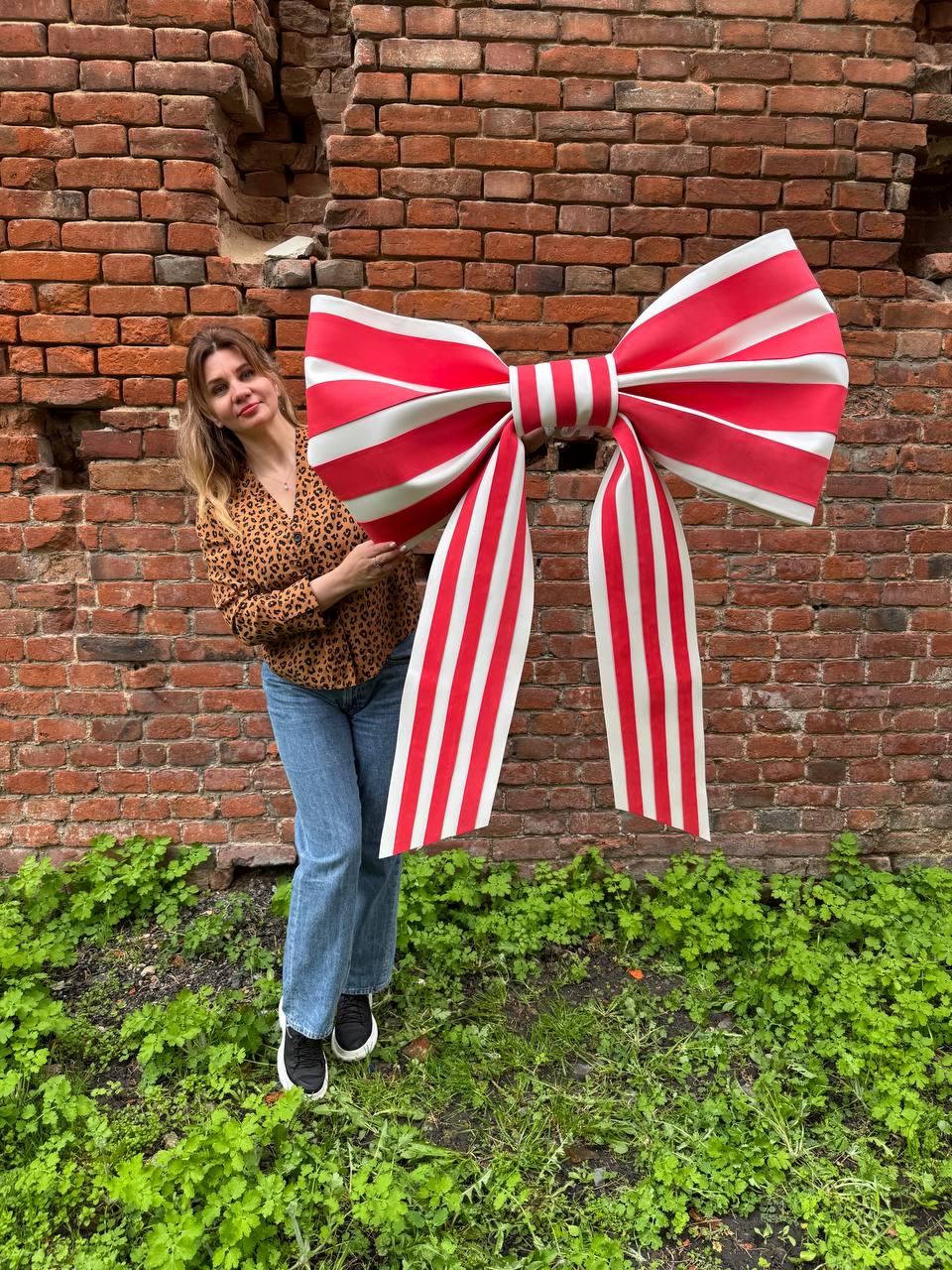 May include: A large, decorative bow with red and white stripes. The bow is held in front of a brick wall and green foliage. The bow's design features a classic bow shape with long, striped streamers.