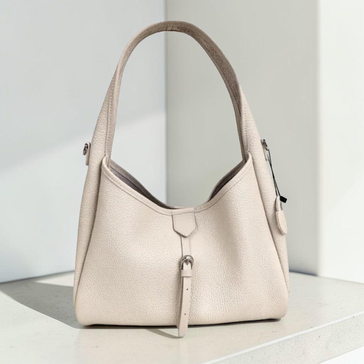 May include: A light beige handbag with a curved top and a single handle. The bag features a small strap with a buckle closure and a zipper detail. The leather bag is sitting on a white surface.