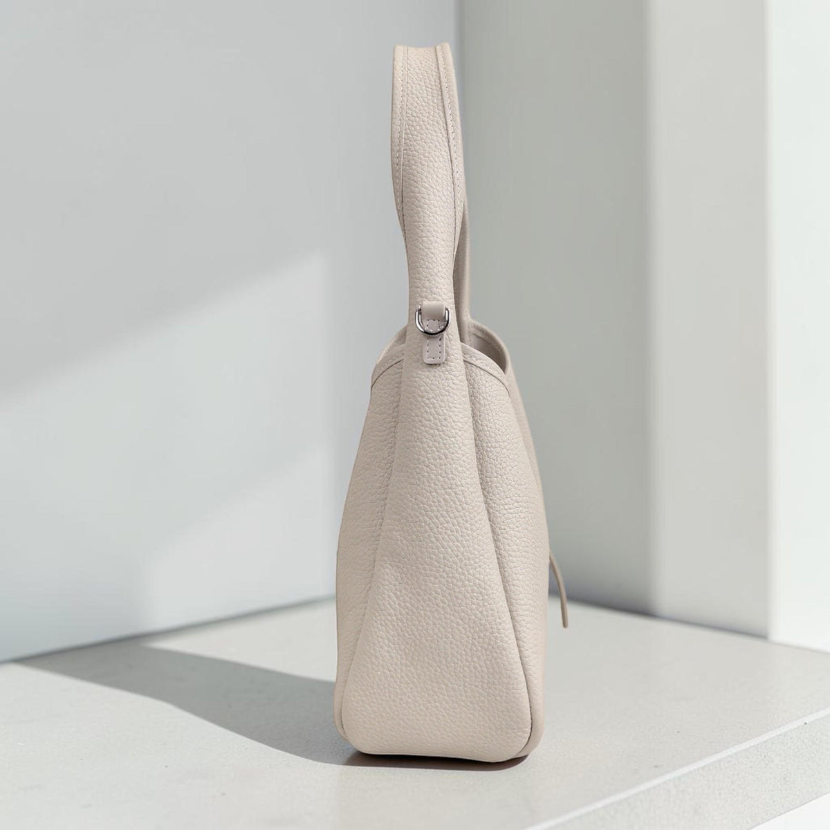 May include: A light beige handbag with a top handle and a shoulder strap attachment. The bag has a textured surface and a rounded bottom. The handle is attached to the bag with a silver-colored metal ring.