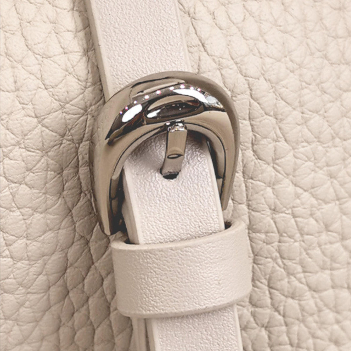May include: Close-up of a cream-colored leather strap with a silver-toned buckle. The buckle is arched and has a shiny finish. The leather has a textured, pebbled appearance.