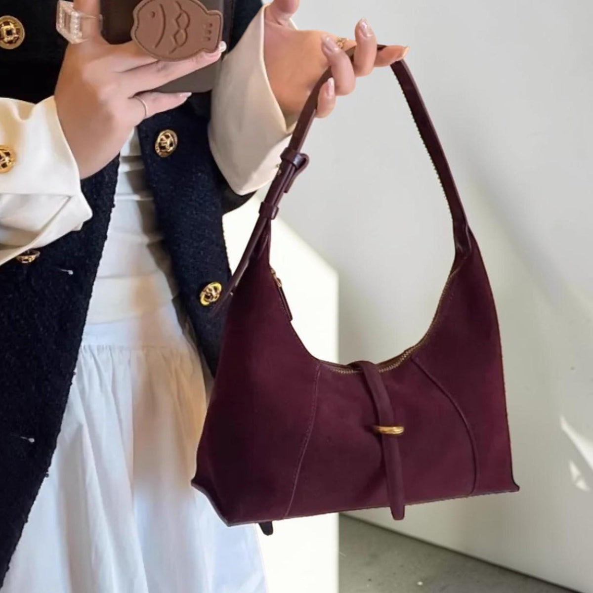 May include: A burgundy suede shoulder bag with a curved design. The bag features a gold-toned zipper and buckle closure. The strap is thin and the bag is held by a person.