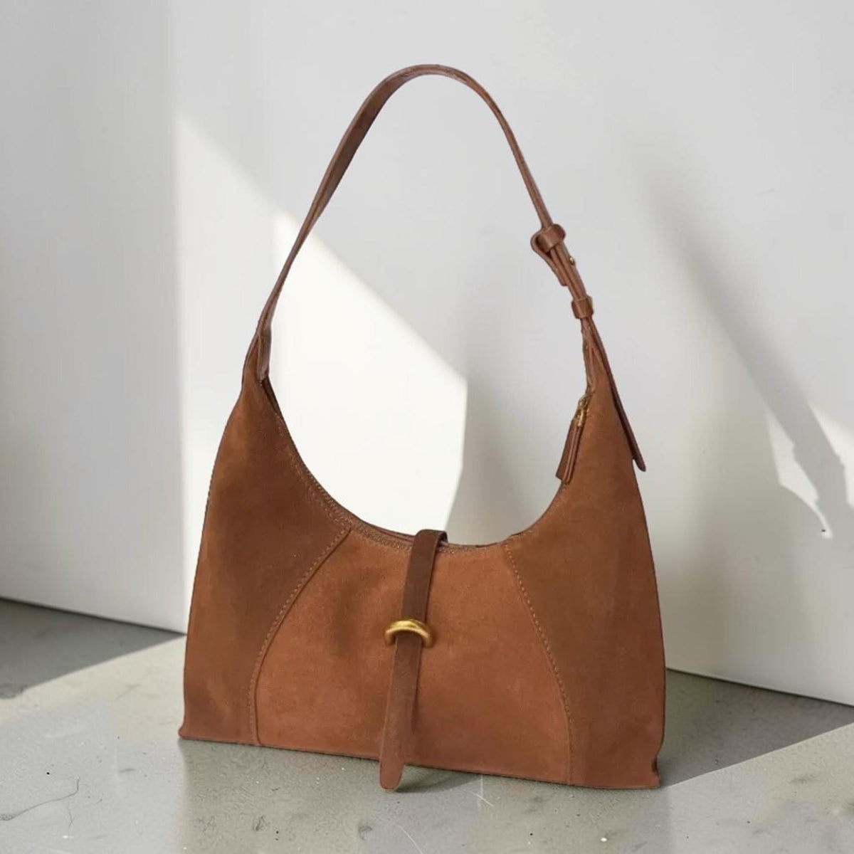 May include: A brown suede shoulder bag with a curved design. The bag features a long, adjustable strap and a small, gold-colored buckle detail. The bag is made of a soft, textured material and has a zipper closure.