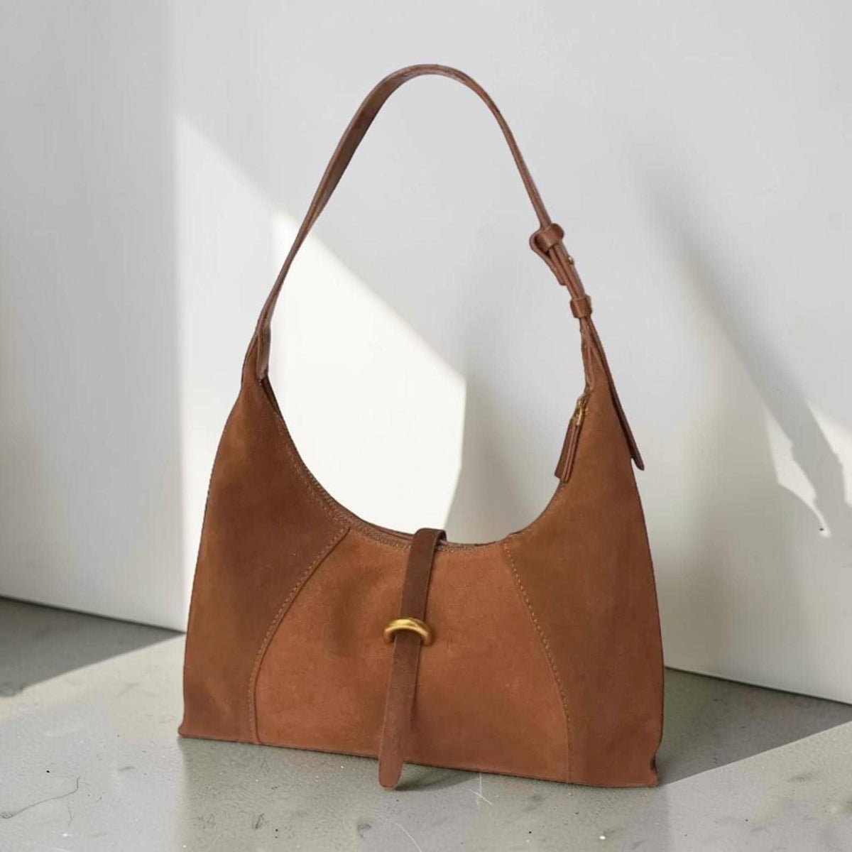 May include: A brown suede shoulder bag with a curved design. The bag features a long, adjustable strap and a small, gold-colored buckle detail. The bag is made of a soft, textured material and has a zipper closure.