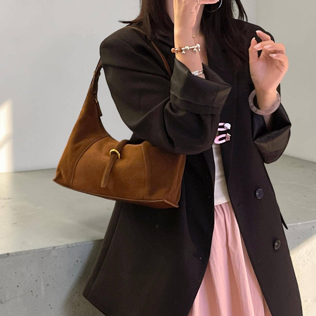 May include: A brown suede shoulder bag with a curved shape and a gold-colored buckle. The bag has a long strap and a small, decorative strap hanging from the buckle. The bag is worn over the shoulder.
