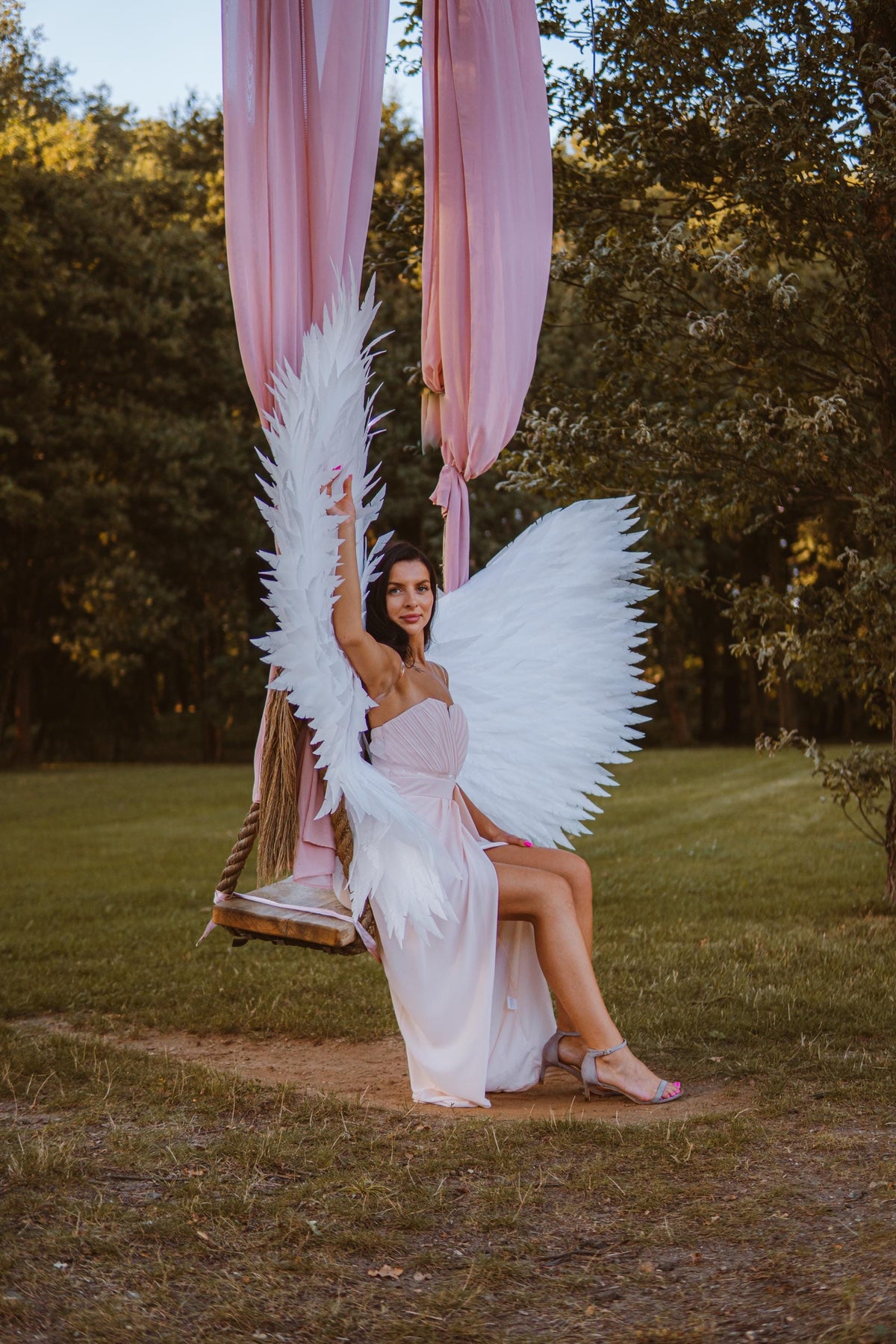 May include: A woman wearing a light pink dress and large white feather angel wings sits on a wooden swing. The swing is suspended from pink fabric draped from above. The background is a grassy area with trees.