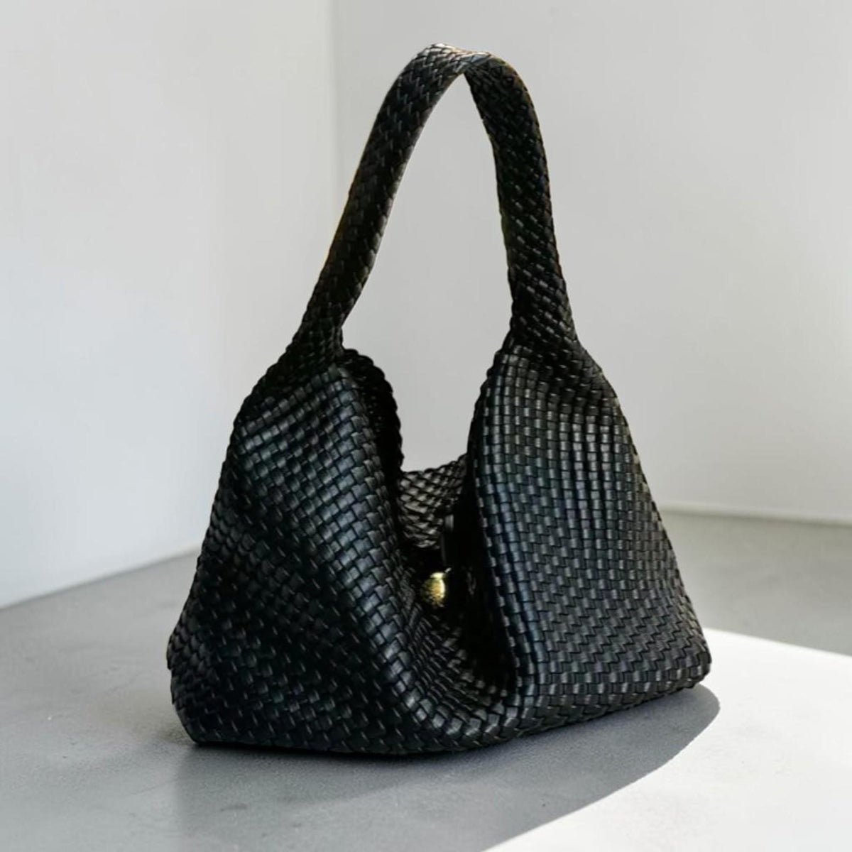 May include: A black woven leather handbag with a single handle. The bag has a rounded shape and a gold-colored clasp. The woven pattern is visible throughout the bag, including the handle. The bag is sitting on a light gray surface.