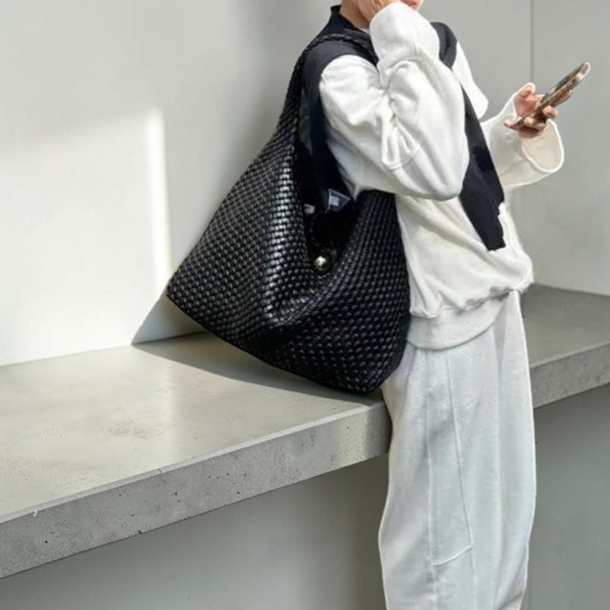 May include: A large black woven tote bag with a shoulder strap. The bag has a rounded shape and a gold-colored button closure. The bag is made of a textured material and appears to be empty.