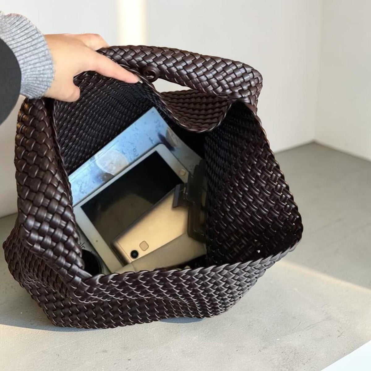 May include: A dark brown woven tote bag with a rectangular shape. The bag is open, revealing a tablet and a smartphone inside. The bag is held open by a person's hand.