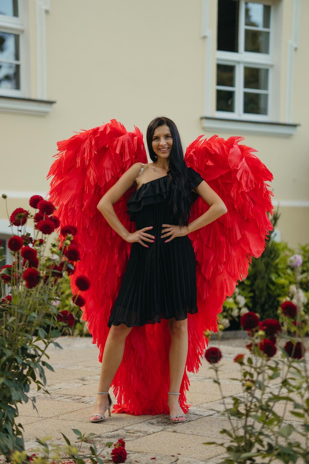 May include: A woman wearing a black, one-shoulder dress and silver heels stands in front of a building. Large, vibrant red angel wings are behind her. The wings are made of layered, textured material. Red flowers and greenery are in the foreground.