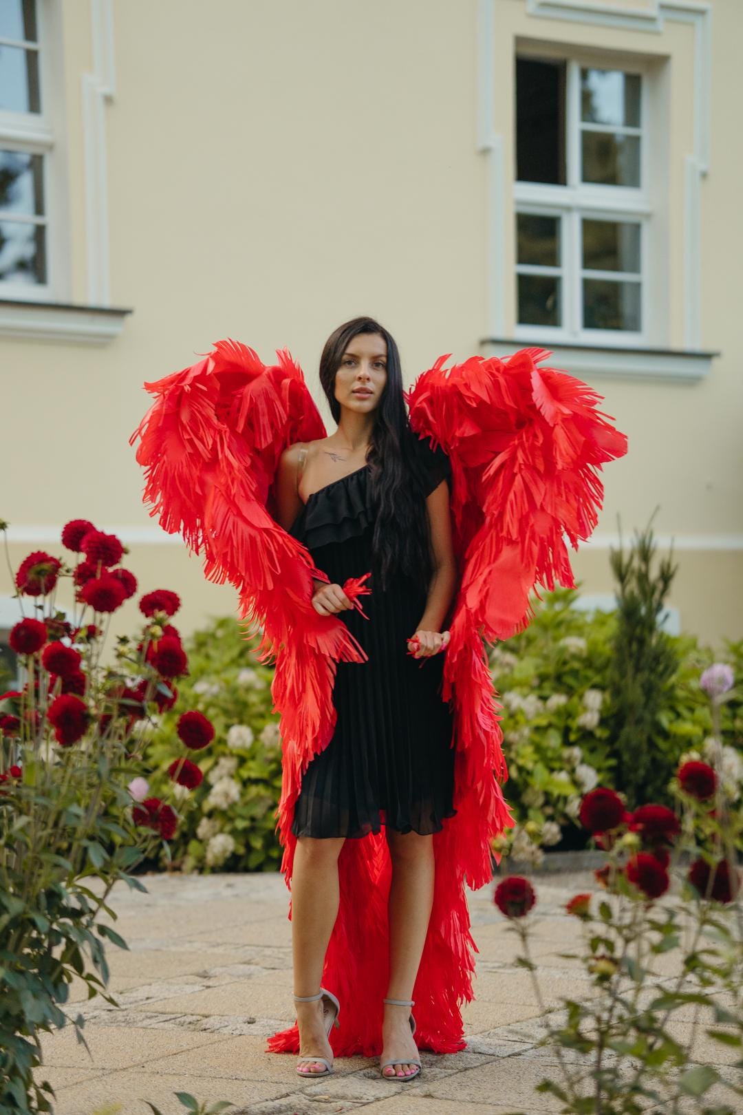 May include: A woman wearing a black, one-shoulder dress and large, vibrant red feather wings. She is standing in a garden with red flowers and a building in the background. The dress is pleated and the wings are full.