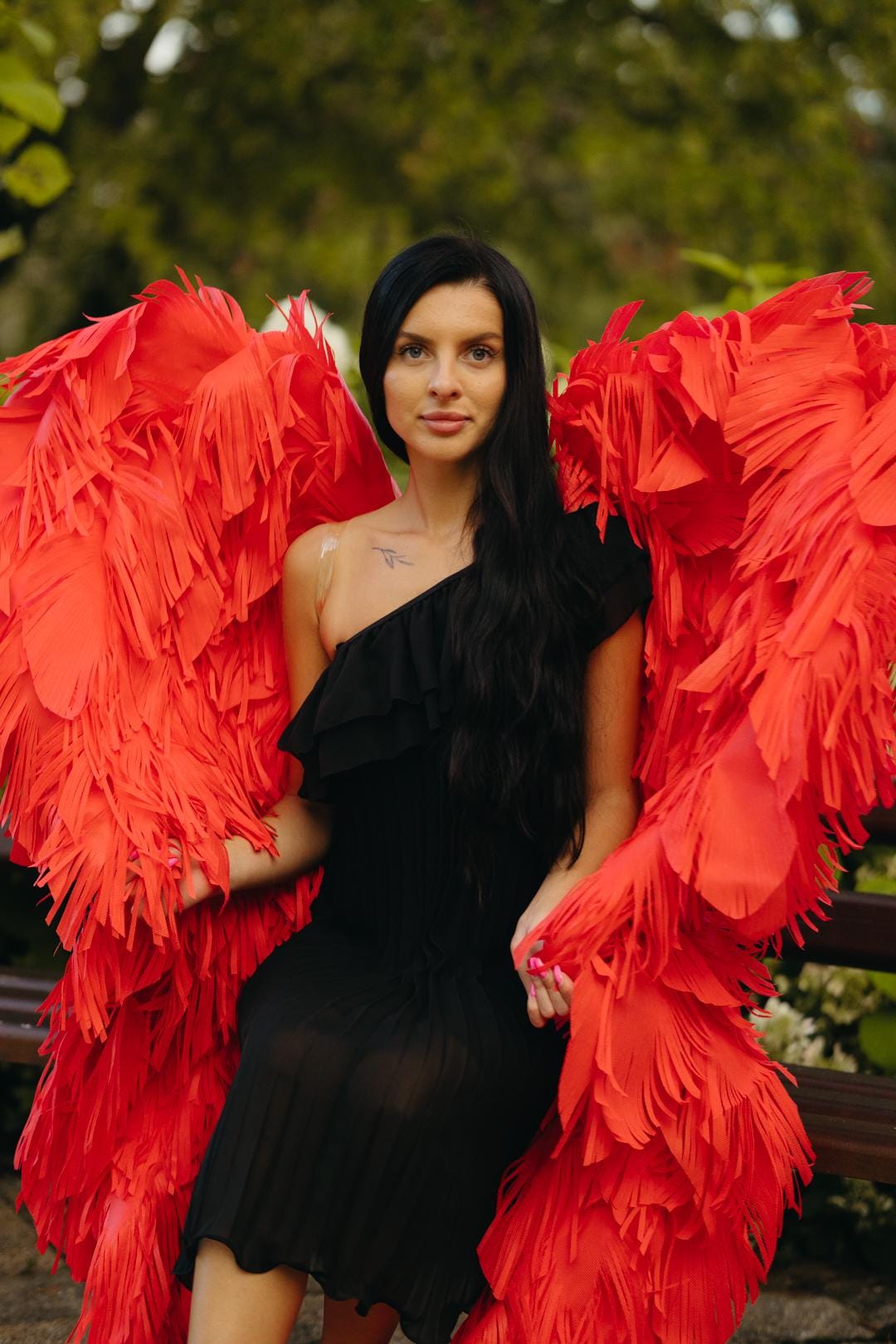 May include: A woman wearing a black, one-shoulder dress and large, vibrant red angel wings. The wings are made of layered, fringed material. The background is a blurred outdoor scene with greenery.