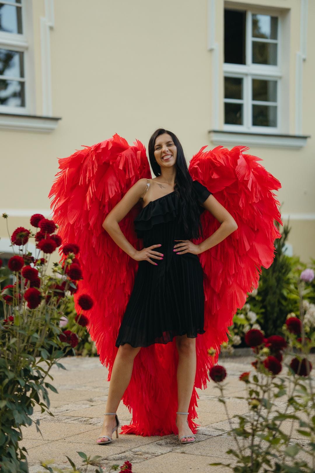 May include: A woman wearing a black one-shoulder dress and red feather wings stands outdoors. The wings are large and heart-shaped. The background includes a building with windows and red flowers.