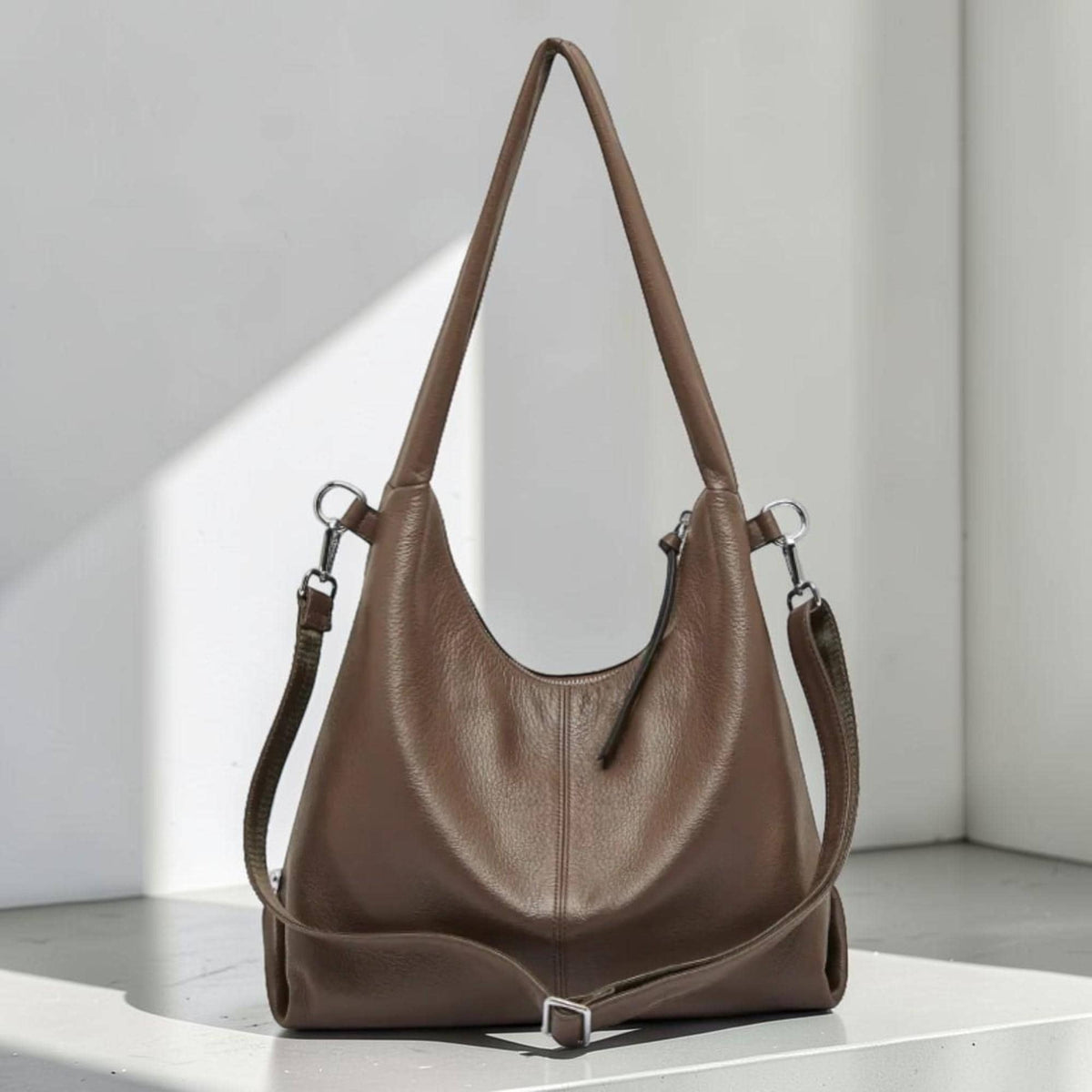 May include: A brown leather shoulder bag with a long strap and a shorter adjustable strap. The bag has a curved shape and a silver-colored buckle. The bag is made of a smooth leather material.