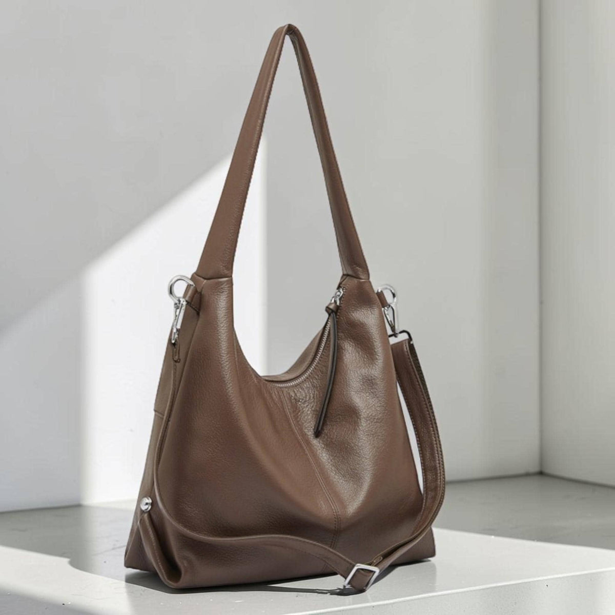 May include: A brown leather shoulder bag with a long strap and a shorter adjustable strap. The bag has a curved shape and a zipper closure. The hardware is silver-colored. The bag is made of a soft, textured leather.