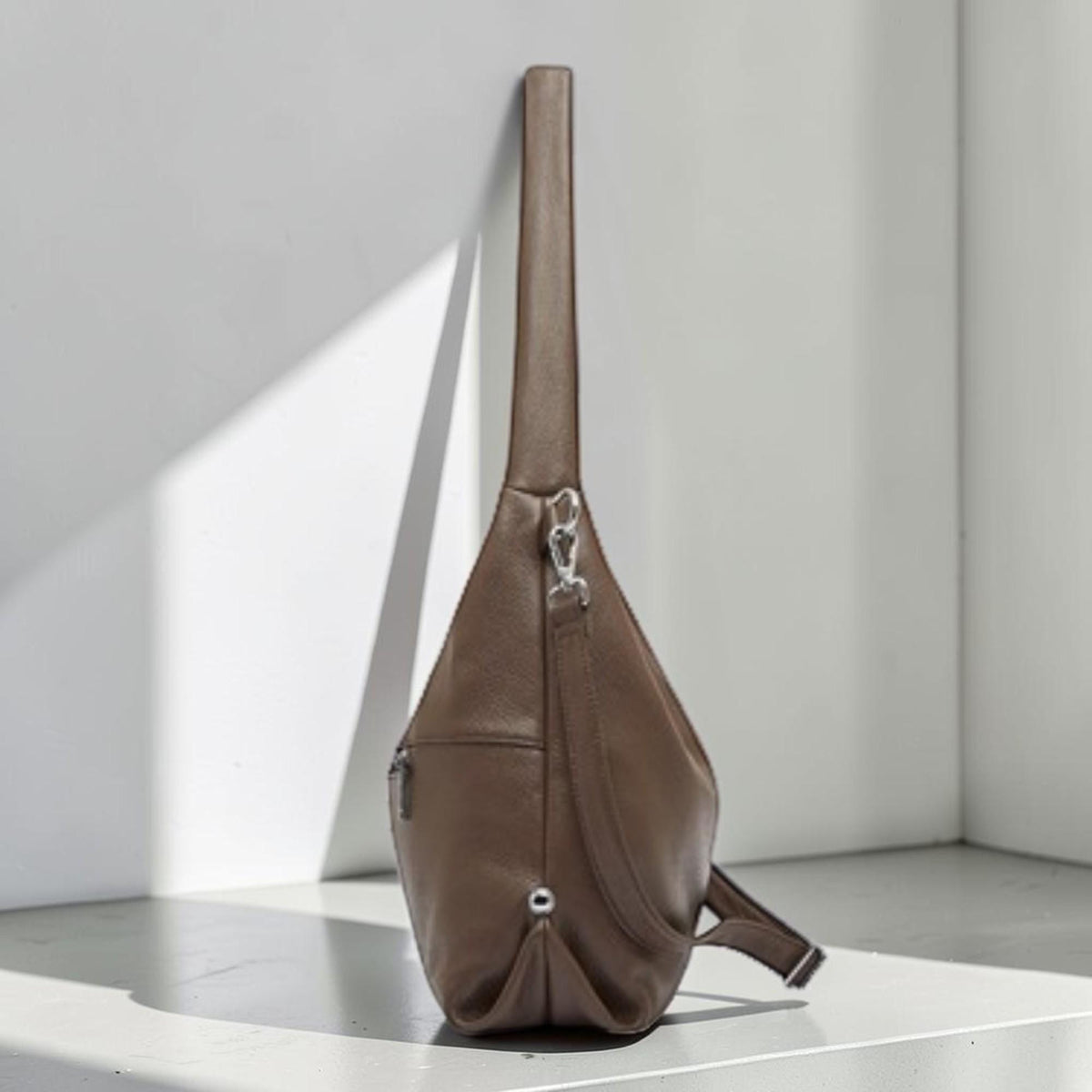 May include: A brown leather sling bag with a long, narrow strap. The bag has a teardrop shape with a zippered pocket and a silver-tone clasp. The bag is leaning against a white wall.
