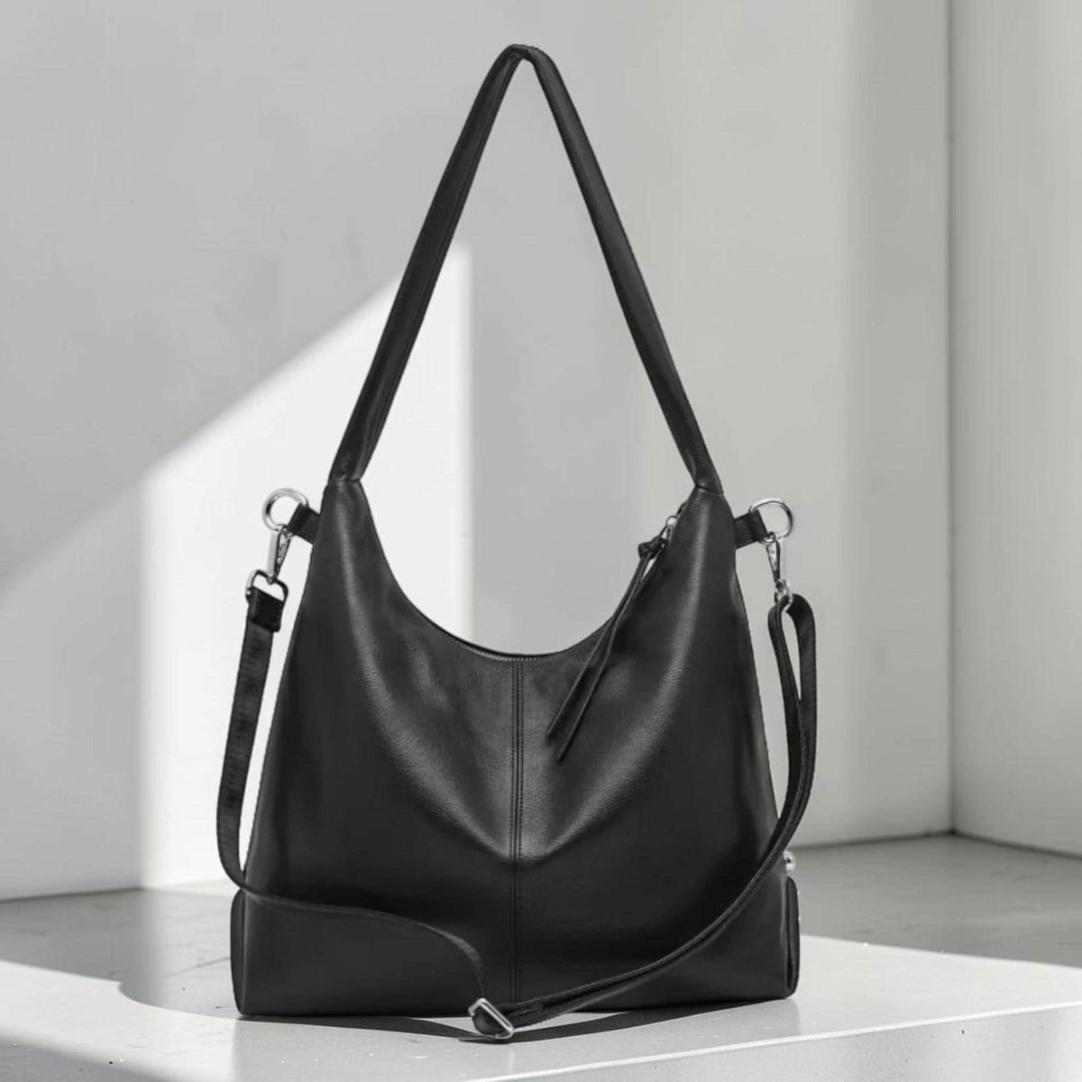May include: A black leather shoulder bag with a long strap and a detachable crossbody strap. The bag has a curved shape and a rectangular base. The bag is made of smooth leather and has silver-tone hardware.