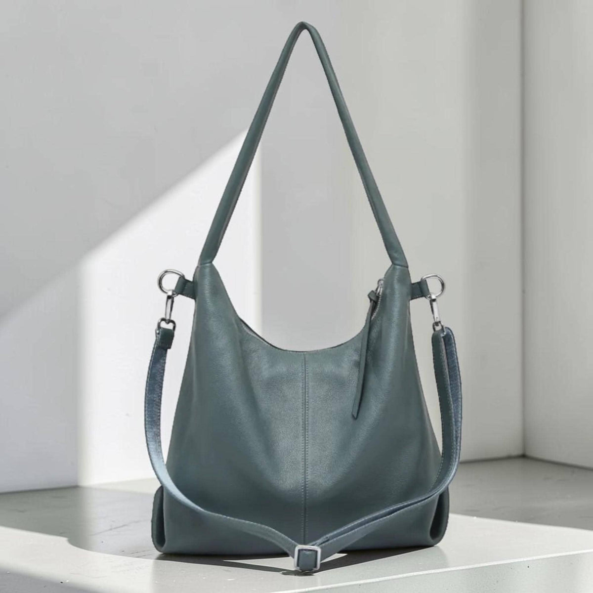 May include: A teal-colored leather shoulder bag with a long adjustable strap. The bag has a curved shape and a silver-tone buckle. The bag is sitting on a white surface.