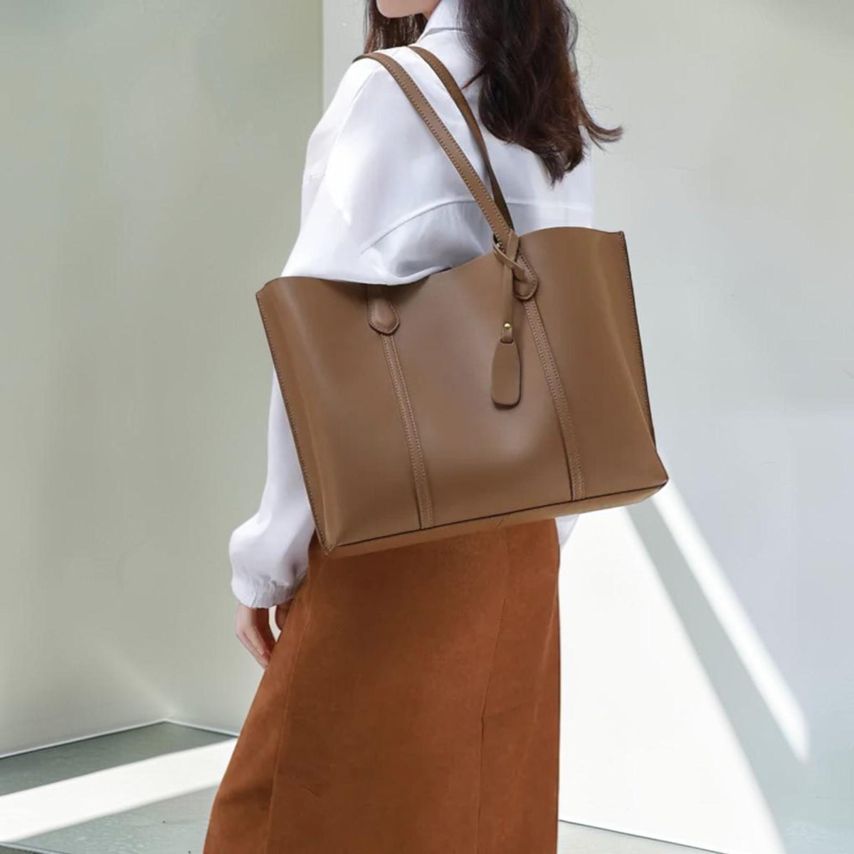 May include: A large, structured brown tote bag with two shoulder straps and a matching hanging leather tag. The bag features a smooth, solid-colored surface with vertical stitched detailing along the front panel and a wide, open-top design.