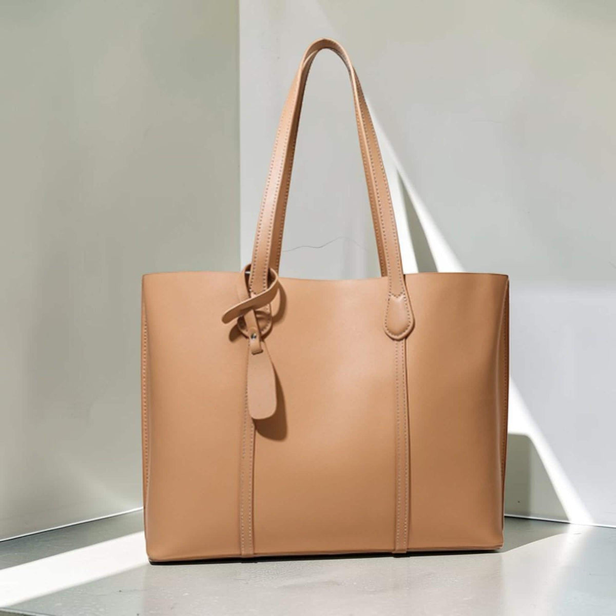 May include: A tan tote bag with a smooth finish, featuring two long shoulder straps, vertical stitched detailing on the front panel, and a small decorative leather tag attached to one of the straps with a metal stud fastener.