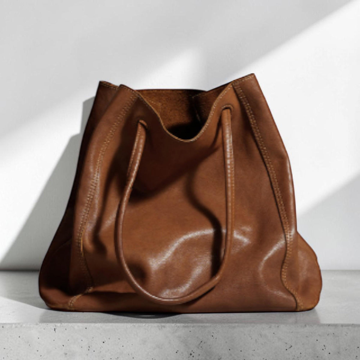 May include: A large, brown leather tote bag with two long handles. The bag has a soft, slouchy shape and a spacious interior. The leather has a natural grain and a warm, earthy tone. The bag is sitting on a gray surface against a white background.