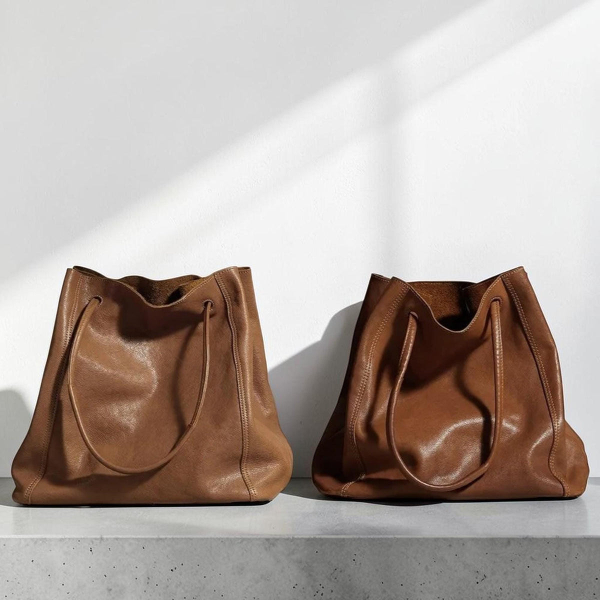 May include: Two brown leather tote bags with drawstring closures. The bags are a warm, medium brown color and have a soft, slouchy appearance. They are sitting on a gray surface against a white wall.