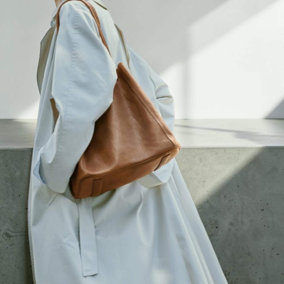 May include: A brown leather tote bag with a shoulder strap. The bag is carried over the shoulder of a person wearing a light blue trench coat. The bag has a trapezoid shape and a simple design.