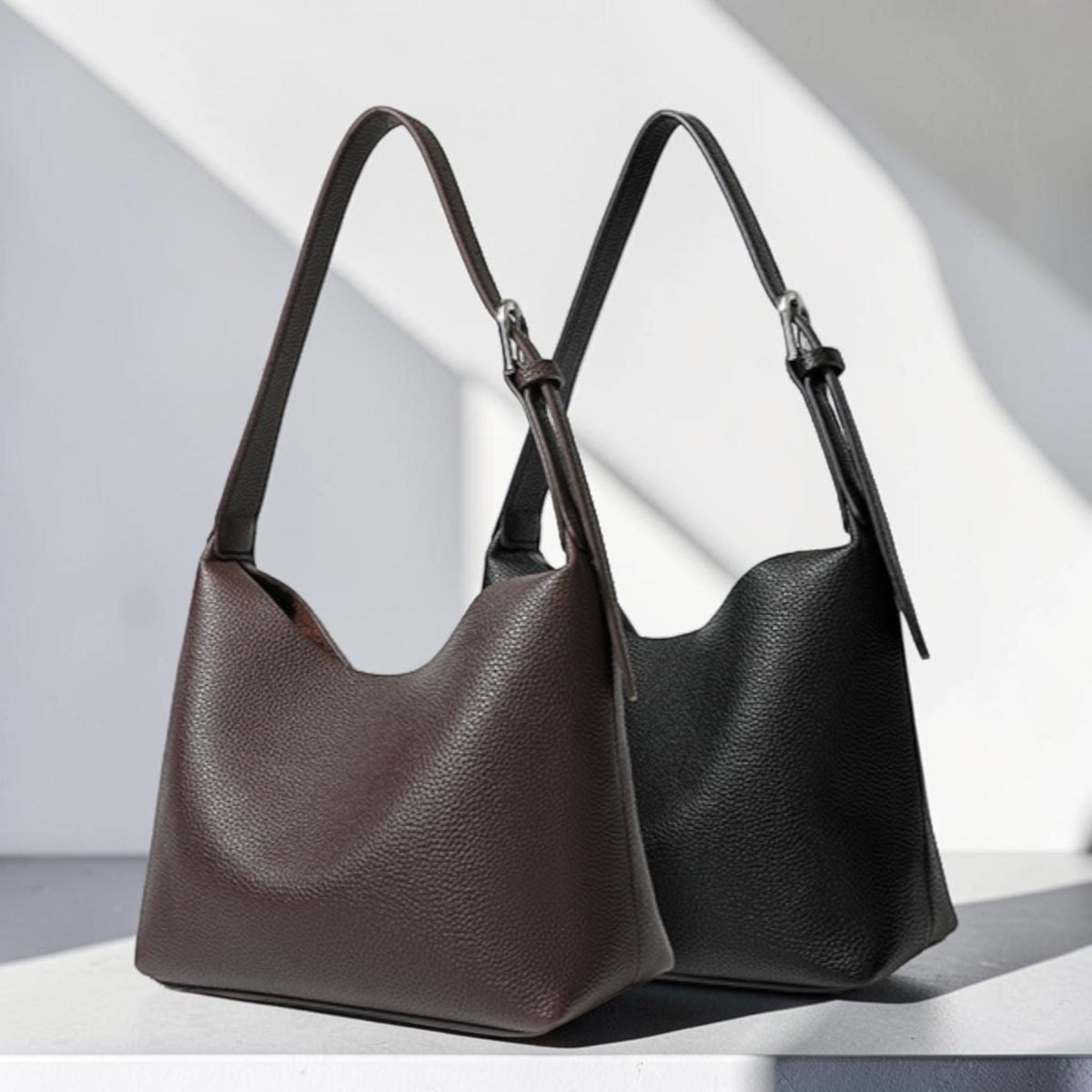 May include: Two leather shoulder bags. One is a deep brown, the other is black. Both bags have a textured surface and long, thin straps with silver-colored buckles. The bags are displayed against a white background with shadows.