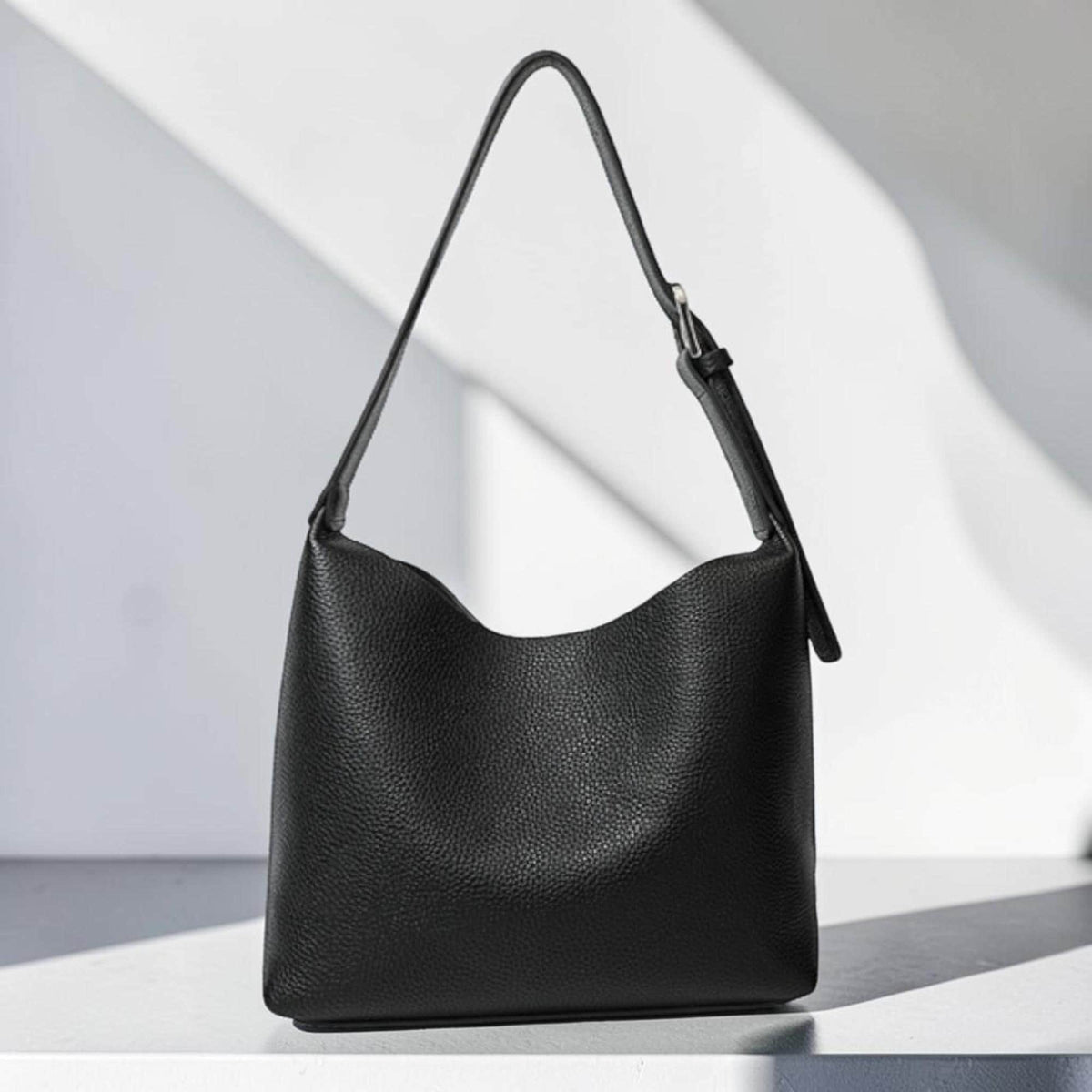 May include: A black leather shoulder bag with a curved top edge and a single adjustable strap. The bag has a simple, minimalist design and a textured surface. The strap is attached with a silver-colored buckle.