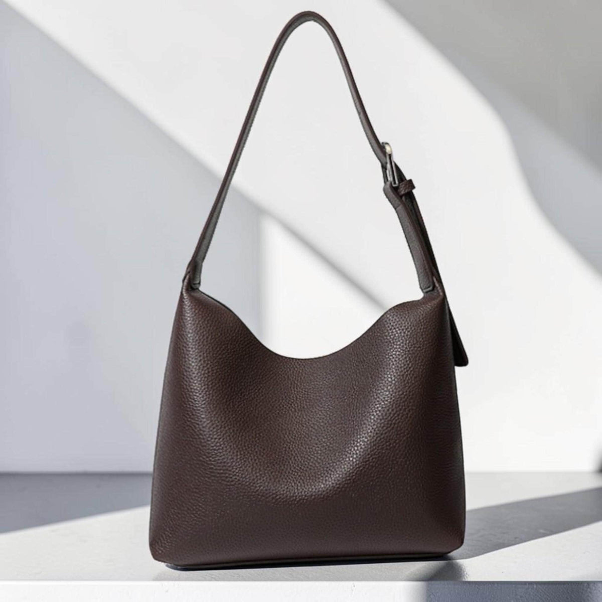 May include: A dark brown leather shoulder bag with a single strap and a curved top. The bag has a textured surface and a silver-colored buckle on the strap. The bag is sitting on a white surface.