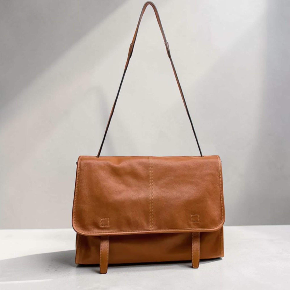 May include: A tan leather messenger bag with a flap closure and a long, thin shoulder strap. The bag has two buckle fastenings on the front and a central vertical seam. The leather appears soft and has a natural grain.