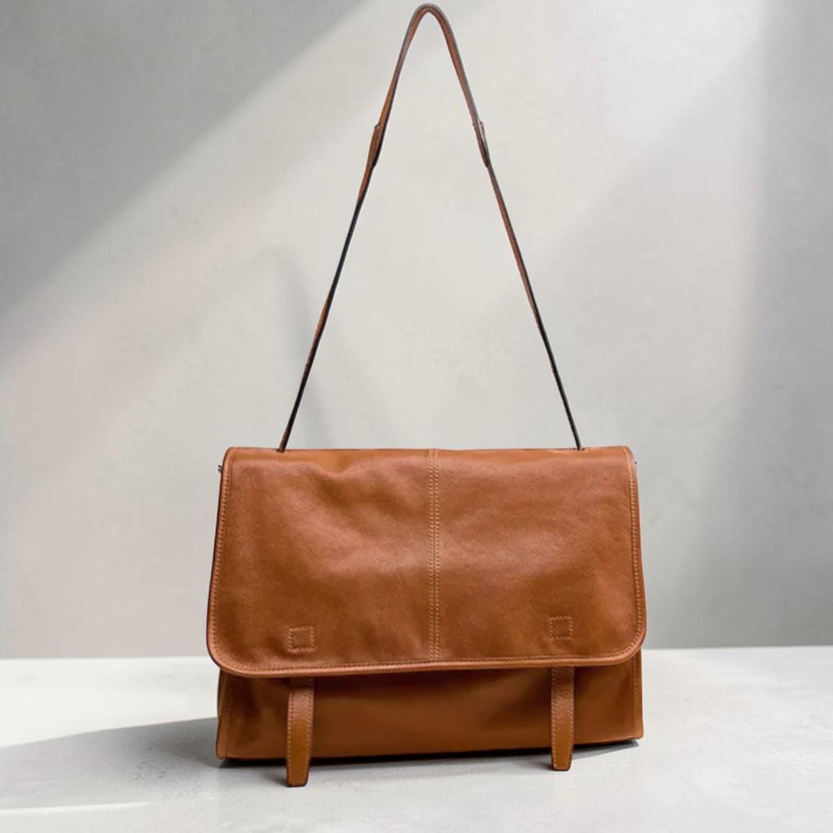 May include: A tan leather messenger bag with a flap closure and a long, thin shoulder strap. The bag has two buckle fastenings on the front and a central vertical seam. The leather appears soft and has a natural grain.